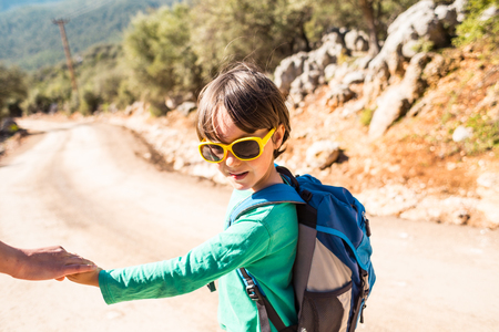 The boy walks with his mother on a mountain road. A woman travels with a child. The kid holds mom's hand. Travel with backpacks. Hike and climb with kids.の写真素材
