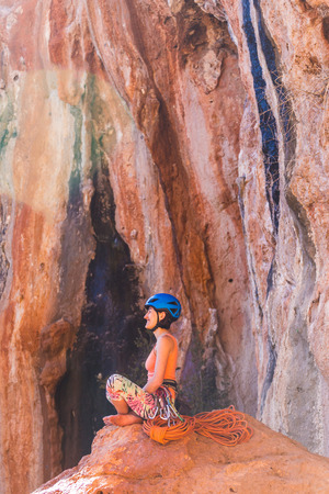 Climber sits near the rope and prepares to overcome the route. Slender girl holds climbing equipment. A woman in a helmet looks at a rock wall. Rest after climbing.の写真素材