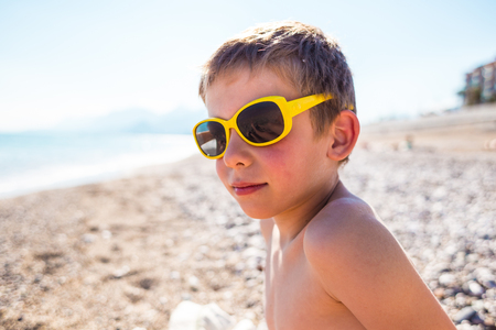 A boy in sunglasses plays on the beach. Portrait of a laughing child. Smiling baby sunbathes on the sea coast. Protect your eyes from sunlight.の写真素材