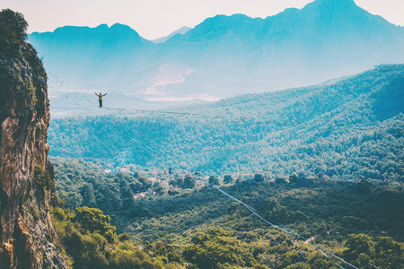 A man is walking along a stretched sling. Highline in the mountains. Man catches balance. Performance of a tightrope walker in nature. Highliner on the background of the mountains.の写真素材