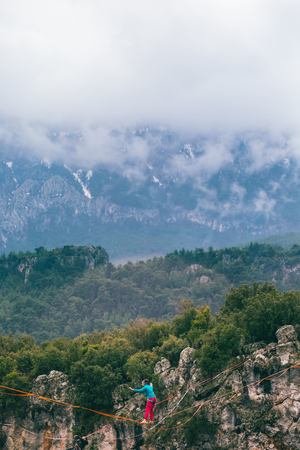 A woman is walking along a stretched sling. Highline in the mountains. Woman catches balance. Performance of a tightrope walker in nature. Highliner on the background of thunderclouds.の写真素材