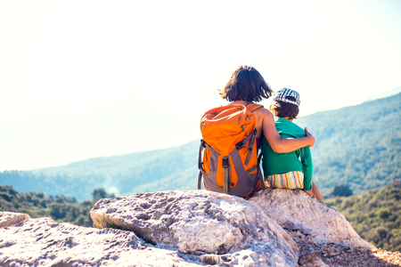 The boy and his mother are sitting on the top of the mountain. A woman travels with a child. Baby hugs mom. Travel with backpacks. Hike and climb with kids. Portrait of a woman with her son.の写真素材