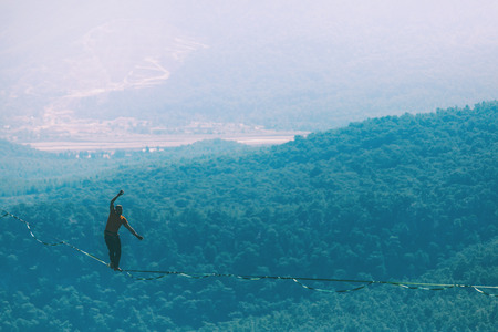 A man is walking along a stretched sling. Highline in the mountains. Man catches balance. Performance of a tightrope walker in nature. Highliner on the background of valley.の写真素材
