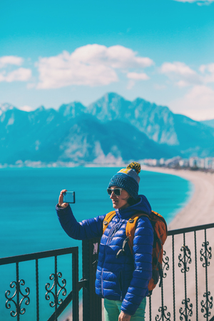 A girl takes a selfie on the waterfront. A woman walks through the resort town and takes pictures. Brunette travels around the Turkish city in winter. Photo on the background of the sea and mountains.の写真素材