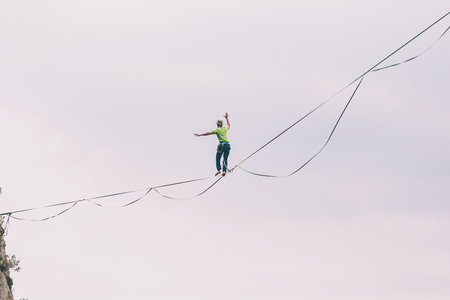Highliner on the background of blue sky makes a move. A man is walking along a stretched sling. Performance tightrope walker. Man balances over the abyss. A decisive step forward.の写真素材
