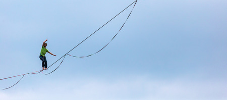 Highliner on the background of blue sky makes a move. A man is walking along a stretched sling. Performance tightrope walker. Man balances over the abyss. A decisive step forward.の写真素材