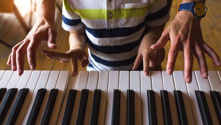 A woman teaches her son to play the piano. The boy masters the keyboard musical instrument. A child learns music. Children's and women's hands on the piano keys. Music lesson. Tutor for a child.の写真素材