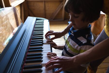 A woman teaches her son to play the piano. The boy masters the keyboard musical instrument. A child learns music. Children's and women's hands on the piano keys. Music teacher. Tutor for a child.の写真素材