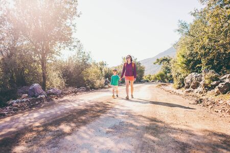 The boy walks with his mother on a mountain road. A woman travels with a child. The kid holds mom's hand. Travel with backpacks. Hike and climb with kids.の写真素材