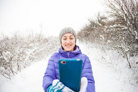 Smartphone. A woman makes a photo on a mobile phone in the winter forest. A smiling girl in a knitted hat makes selfie against the background of a snow covered park.の写真素材
