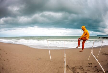A man sits on the beach on a windy day. Sea coast in the cold season. Ocean waves and overcast sky. Watching the sea alone. A man in bright clothes is looking at the water.の写真素材