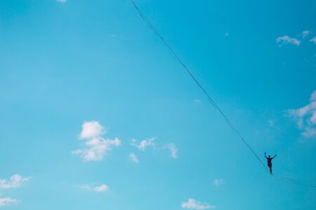 Highliner on the background of blue sky makes a move. A man is walking along a stretched sling. Performance tightrope walker. Man balances over the abyss. A decisive step forward.の写真素材