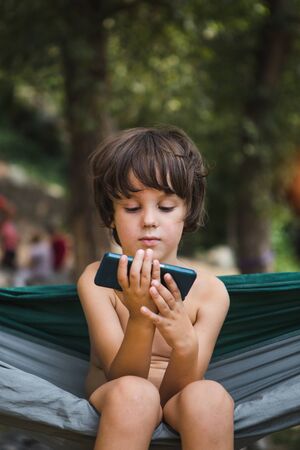 A child sits in a hammock and looks into a smartphone. A boy is playing on the phone. A child reads while resting.の写真素材