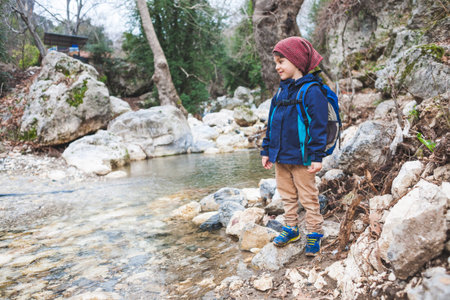 A child with a backpack stands near a mountain river. The boy looks at the creek. The kid walks...