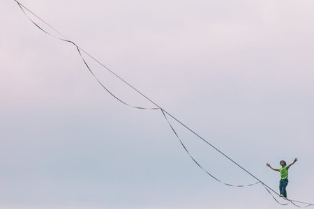 Highliner on the background of blue sky makes a move. A man is walking along a stretched sling. Performance tightrope walker. Man balances over the abyss. A decisive step forward.の写真素材