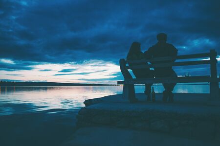 A couple sits on a bench and looks at the sea. A man and a woman on a date. Lovers drink wine on the beach at sunset. Bench on the pier.の写真素材