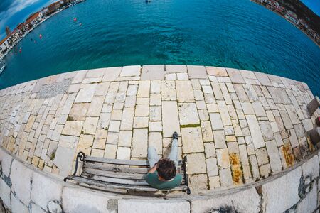 A young man sits on a bench and looks at the water. Walk along the promenade. A man is waiting for someone and looking at the sea. Time spent alone. Embankment in a small Croatian town.の写真素材