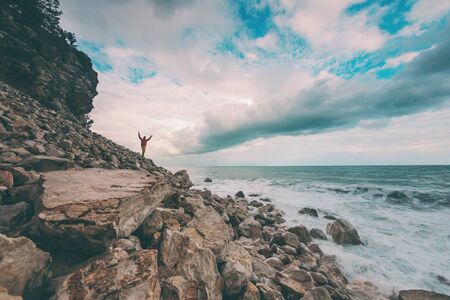 A man stands a huge stone against the sea and a beautiful sky. Traveler walks along the coast of the ocean. Storm waves and beautiful clouds. The man raised his hands up.の写真素材