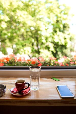 Mobile phone and a cup of coffee on the windowsill. Glass cup with latte near the window. A cozy coffee shop.の写真素材