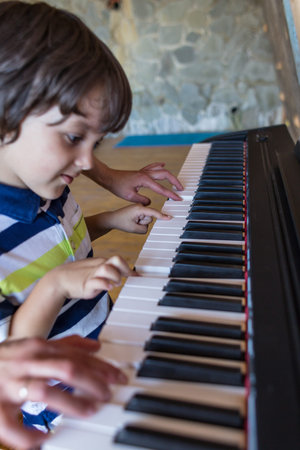 A woman teaches her son to play the piano. The boy masters the keyboard musical instrument. A child learns music. Children's and women's hands on the piano keys. Music lesson. Tutor for a child.の写真素材