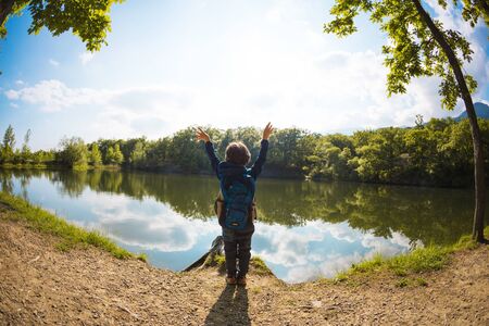 A child on the lake. A boy with a backpack stands on the banks of the river. The kid walks through the woods. Walk in the park on a sunny spring day. The boy looks at the water.の写真素材