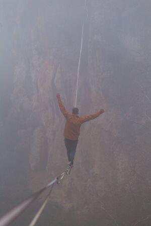 A man is walking along a stretched sling in the fog. Highline in the mountains. Man catches balance. Performance of a tightrope walker in nature. Highliner on the background of thunderclouds.の写真素材