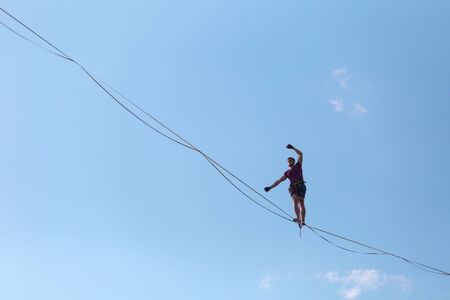 Highliner on the background of blue sky makes a move. A man is walking along a stretched sling. Performance tightrope walker. Man balances over the abyss. A decisive step forward.の写真素材