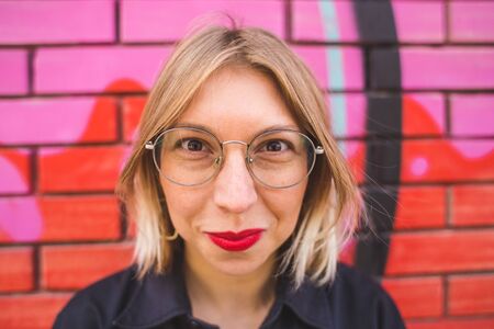 Portrait of a blonde in glasses on a brick wall background. A woman walks around the city. The smiling girl. Lips with red lipstick.の写真素材