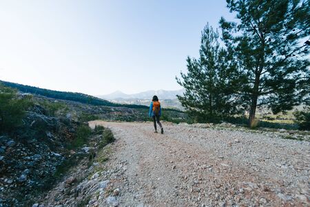 A woman with a backpack is walking along a mountain road. Girl on a background of beautiful mountains. Traveling to scenic spots. Hiking alone.の写真素材