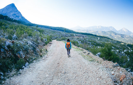 A woman with a backpack is walking along a mountain road. Girl on a background of beautiful mountains. Traveling to scenic spots. Hiking alone.の写真素材