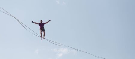 Highliner on the background of blue sky makes a move. A man is walking along a stretched sling. Performance tightrope walker. Man balances over the abyss. A decisive step forward.の写真素材
