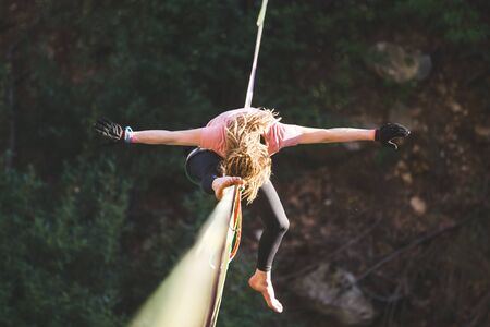 A woman is sitting on a highline, Highliner on a stretched sling, The girl does stretching on a sling stretched over the forest, Trying to get up, highlining, A tightrope walker catches a balance.の写真素材