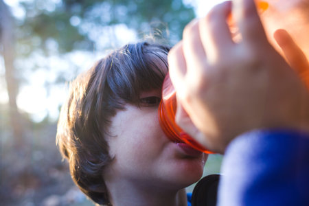 A child drinks water from a refillable bottle, The boy quenches thirst after a walk in the park,...