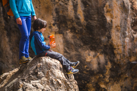 A child with a backpack drinks water from a refillable bottle. The boy sits on a large stone and quenches his thirst. Traveling with children. Woman with a child hiking in the mountains.の写真素材