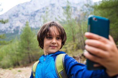 A boy with a backpack makes selfie on a smartphone on a background of beautiful mountains. Child on a camping trip. The kid uses a mobile phone.の写真素材