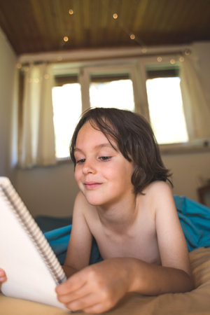 The boy lies in bed and reads a book, Entertainment during quarantine, The child holds a notebook in his hands, Portrait of a smiling boy.の写真素材