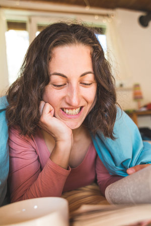 Smiling girl reads a book and drinks coffee while lying in bed. Morning tea. Quarantined time. A woman is engaged in self-development in quarantine.の写真素材