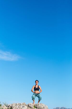 Girl practices yoga on top of the mountain, woman performs asanas standing on a large stone against a background of mountains and a sky, Meditation in nature, Exercise for the development of balance.の写真素材