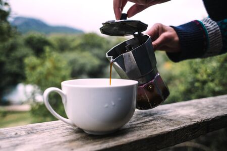 A woman pours coffee into a white cup from a geyser coffee maker, Morning hot drink in nature, Girl is making coffee on the porch of a wooden house.の写真素材