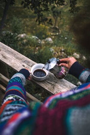 A woman pours coffee into a white cup from a geyser coffee maker, Morning hot drink in nature, Girl is making coffee on the porch of a wooden house.の写真素材