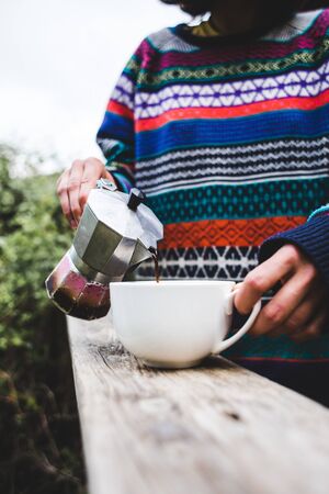 A woman pours coffee into a white cup from a geyser coffee maker, Morning hot drink in nature, Girl is making coffee on the porch of a wooden house.の写真素材