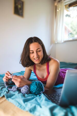 A woman is learning to knit on video lessons from the Internet, The development of new skills, Knitting, Girl knits while sitting on a sofa and watching a movie on a laptop, Hobbies during quarantine.の写真素材