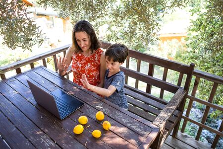 Mom with a child sits on the terrace of a wooden house and look at the laptop. A boy with his mother communicates with relatives on the Internet during quarantine. A woman with son in the courtyard.の写真素材
