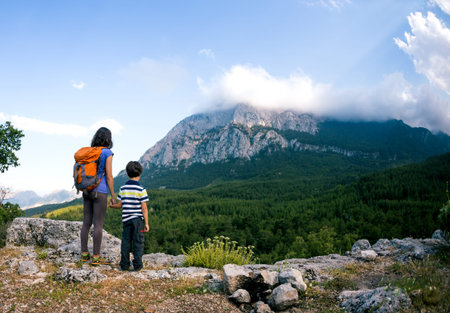 The boy and his mother are standing on the top of the mountain, A woman is traveling with child, Boy with his mother looking at the mountains, Travel with backpacks, Hike and climb with kids.の写真素材