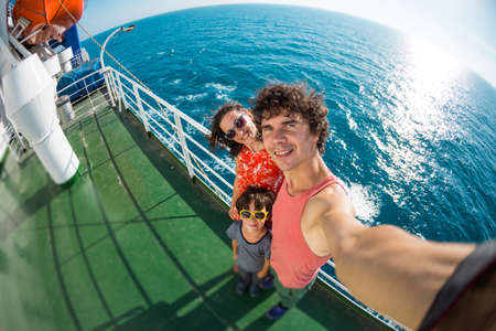 The family is sailing on a cruise ship, a girl with husband and son are standing at the fence on the ship and looking at the sea, traveling by ferry, a boy with mom and dad take selfies on the ocean.の写真素材