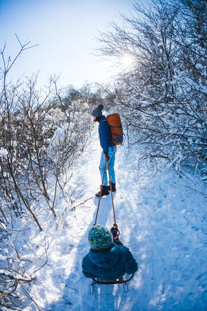 A woman pulls a sled with a child. Mother walks with her son through a snowy forest. Winter walk in the park. The boy rides on a sled. Winter activities with children.の写真素材