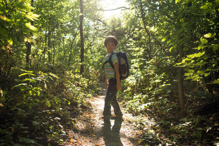 A boy with a backpack walks through the forest, a child explores wildlife, a kid stands alone among the trees, a portrait of a boy.の写真素材
