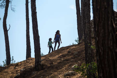 A woman walks with her son through the forest, The boy with his mother go hiking, Silhouette of a woman with backpack and a child against the sky, Travel with children, Mountain trail, Mothers Day.の写真素材
