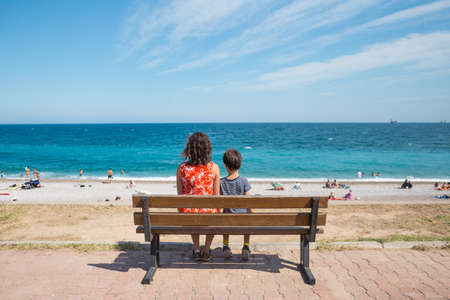 A woman with a child is sitting on a bench on the embankment and looking at the sea, a boy with his mother on a walk along the sea, rest and travel with children, vacation in Turkey.の写真素材
