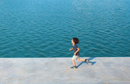 A child runs along the promenade along the water. The boy goes in for sports. Toddler having fun on the street. Doing sports in the city.の写真素材